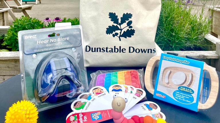 a cotton drawstring bag with the National Trust logo and Dunstable Downs printed on it. Sits on a table surrounded by sensory items: earmuffs, fidget toys, feelings fan and easy hold wooden magnifier.